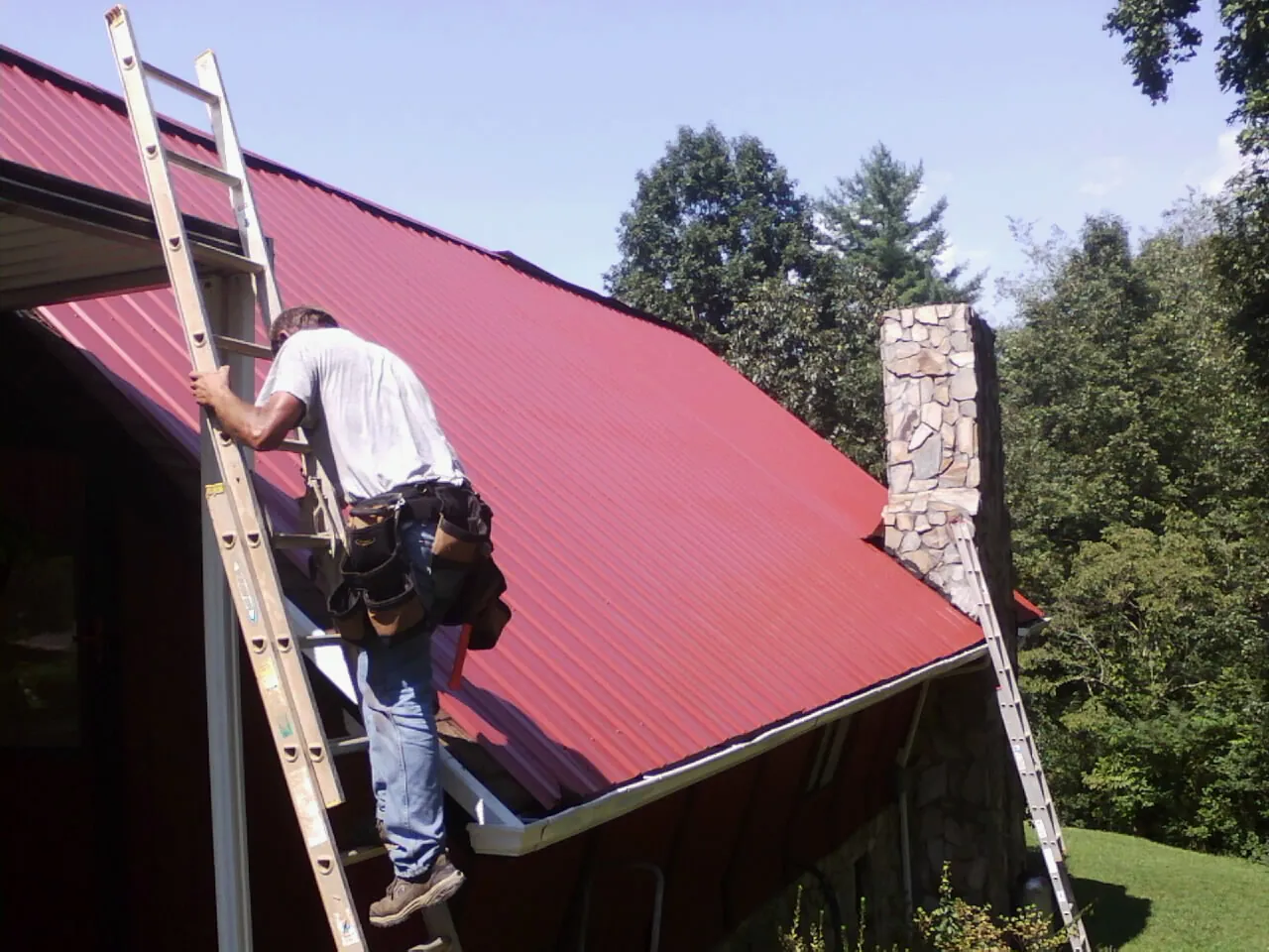Roofer on a ladder beside a red metal roof and stone chimney — Soffit & Fascia Repair by Roofing Brothers in Tatamy